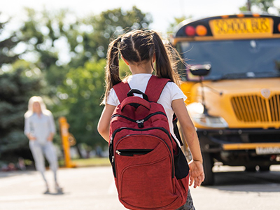 A young child in pigtails, wearing a red backpack, walks toward a yellow school bus on a sunny day. An adult stands in the background near a tree-lined area.