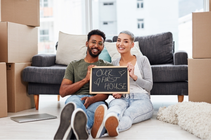 couple sit among moving boxes with a sign that says Our First Home