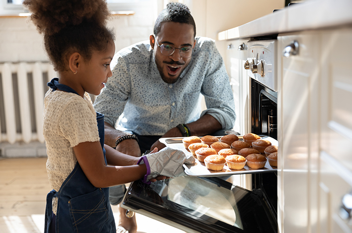 father supervises young daughter taking cupcakes from oven